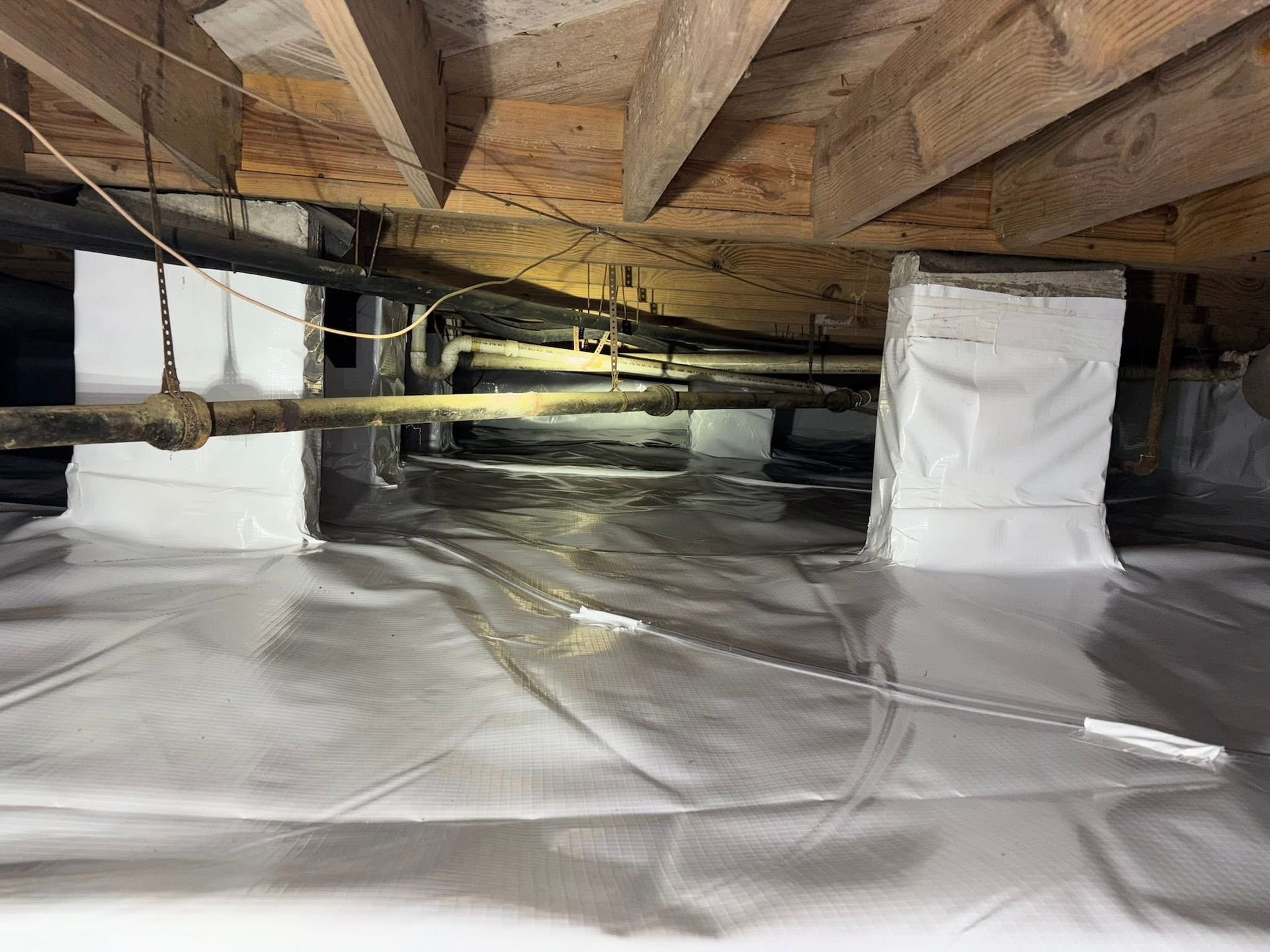 A crawl space with a vapor barrier covering the floor and white insulation wrapped around support columns and walls.