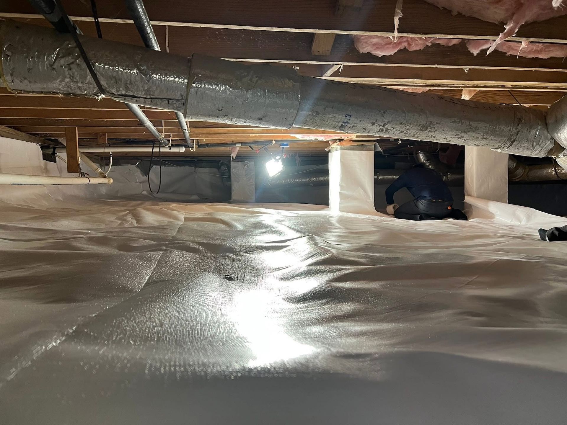 A person crawls under a house in a crawl space encapsulated with white vapor barrier plastic and exposed wooden beams.