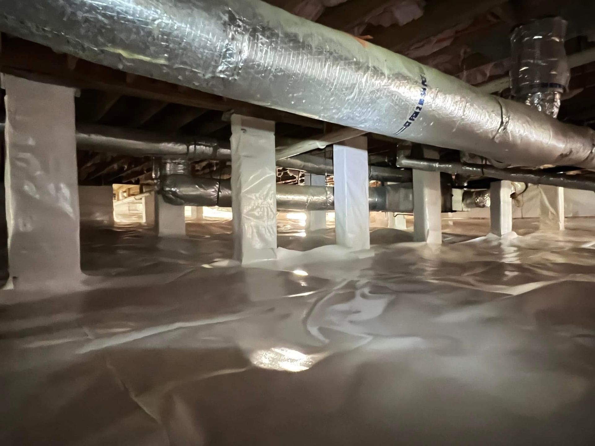 Crawl space with white vapor barrier flooring and wrapped support pillars, featuring insulated metallic HVAC ductwork.