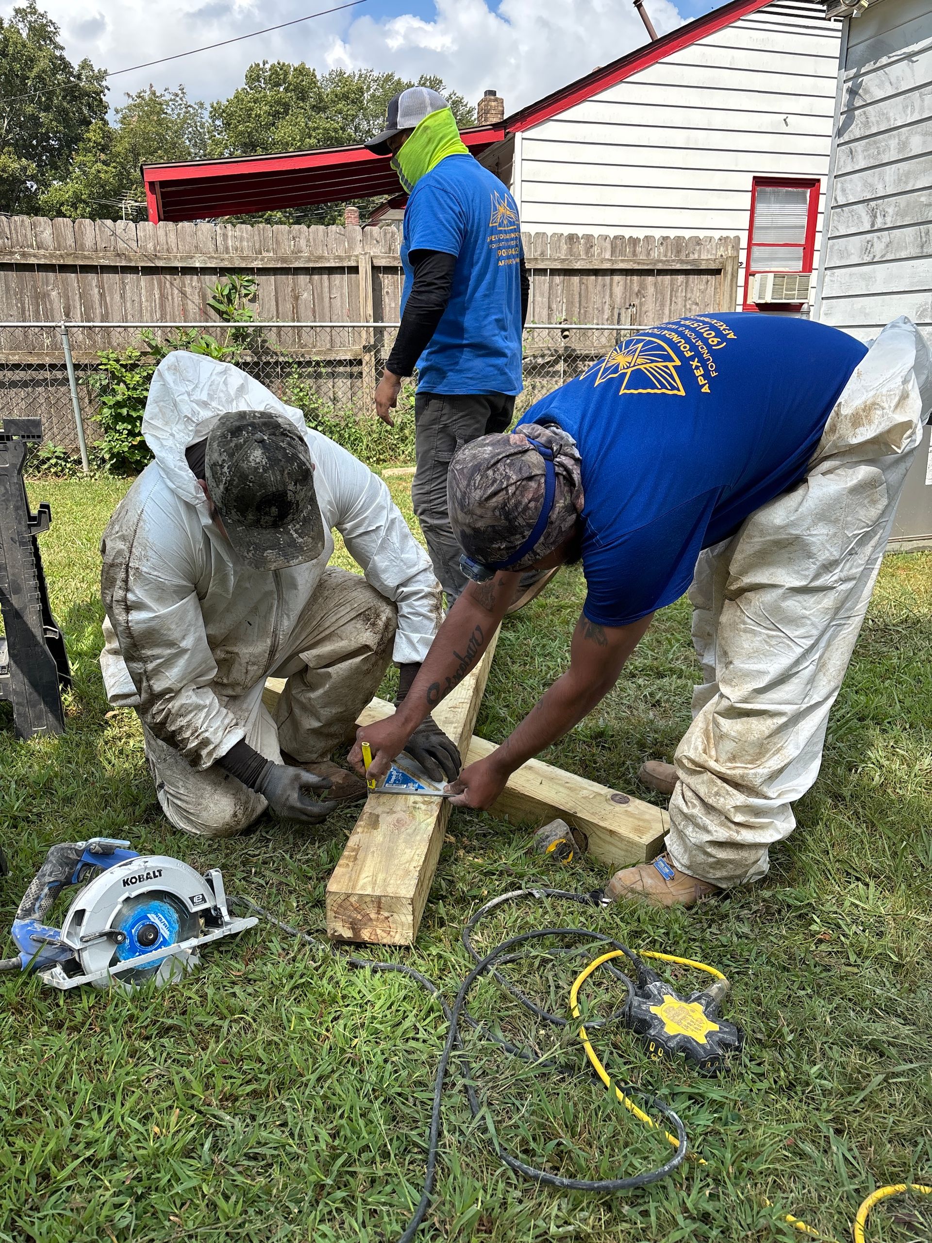 Three workers in protective gear and blue shirts collaborate on a wooden construction project in a grassy yard.