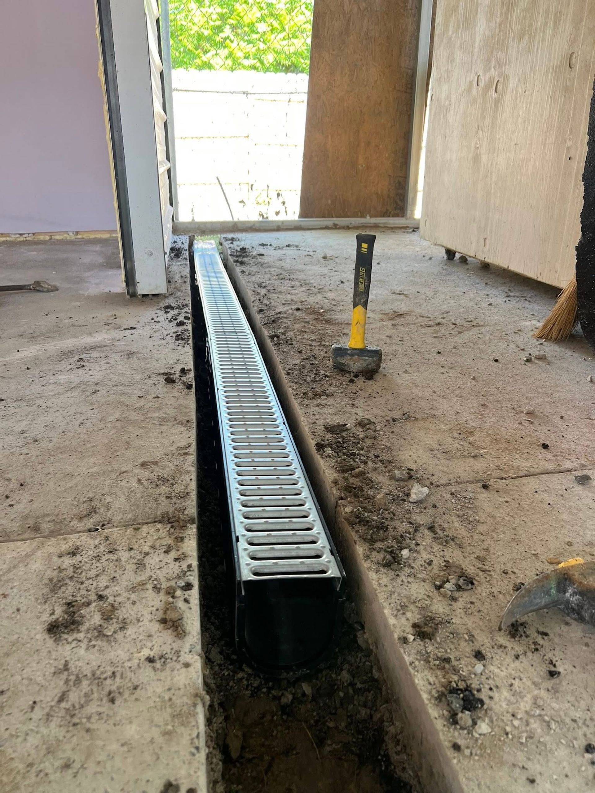 A linear drainage channel installed in a trench in a concrete floor, with a hammer resting on the nearby surface.