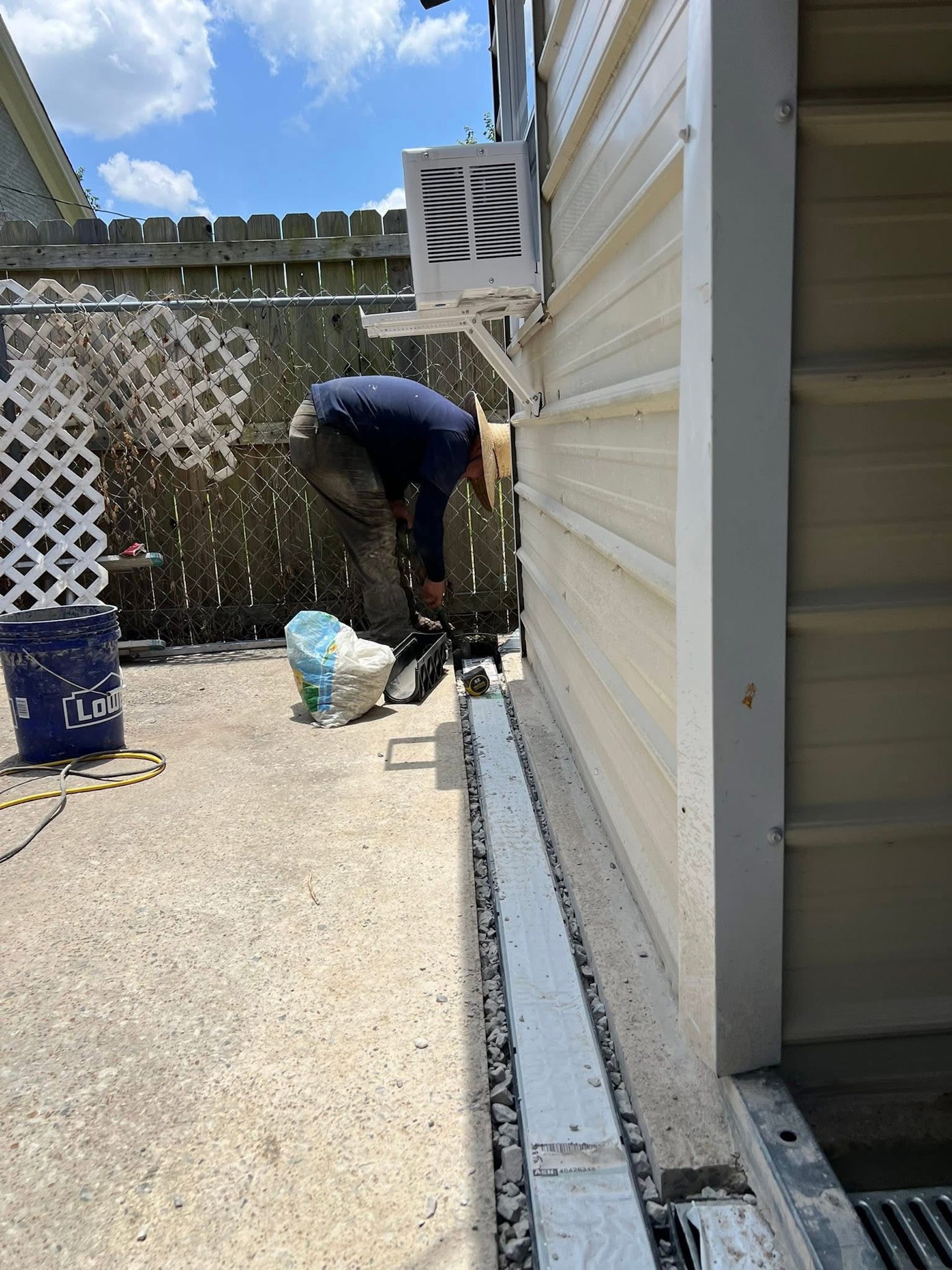A person wearing a wide-brimmed hat works on a gravel trench along the base of a light-colored building wall.