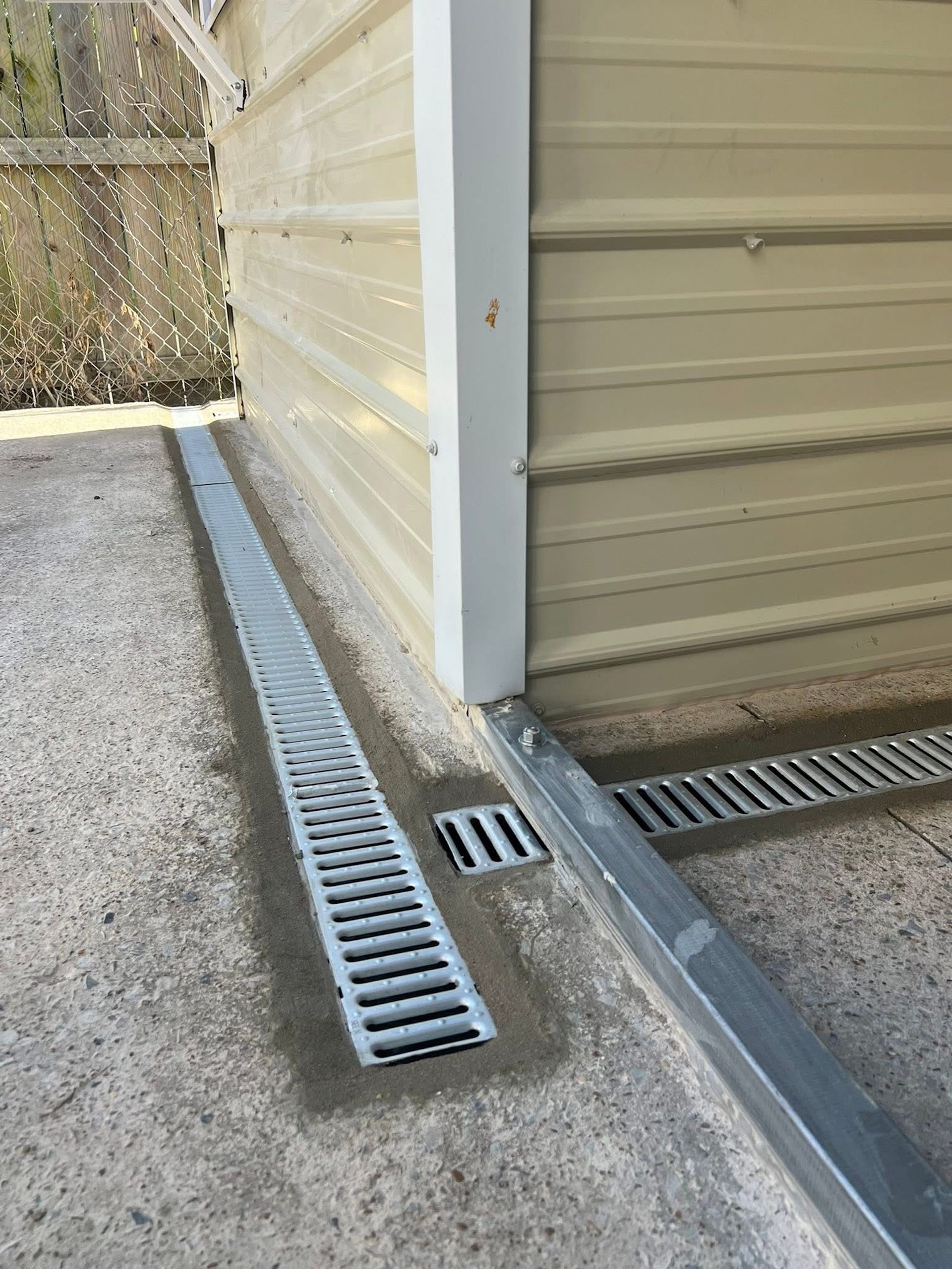 A beige building exterior with a metal channel drain installed in the concrete ground along its foundation.