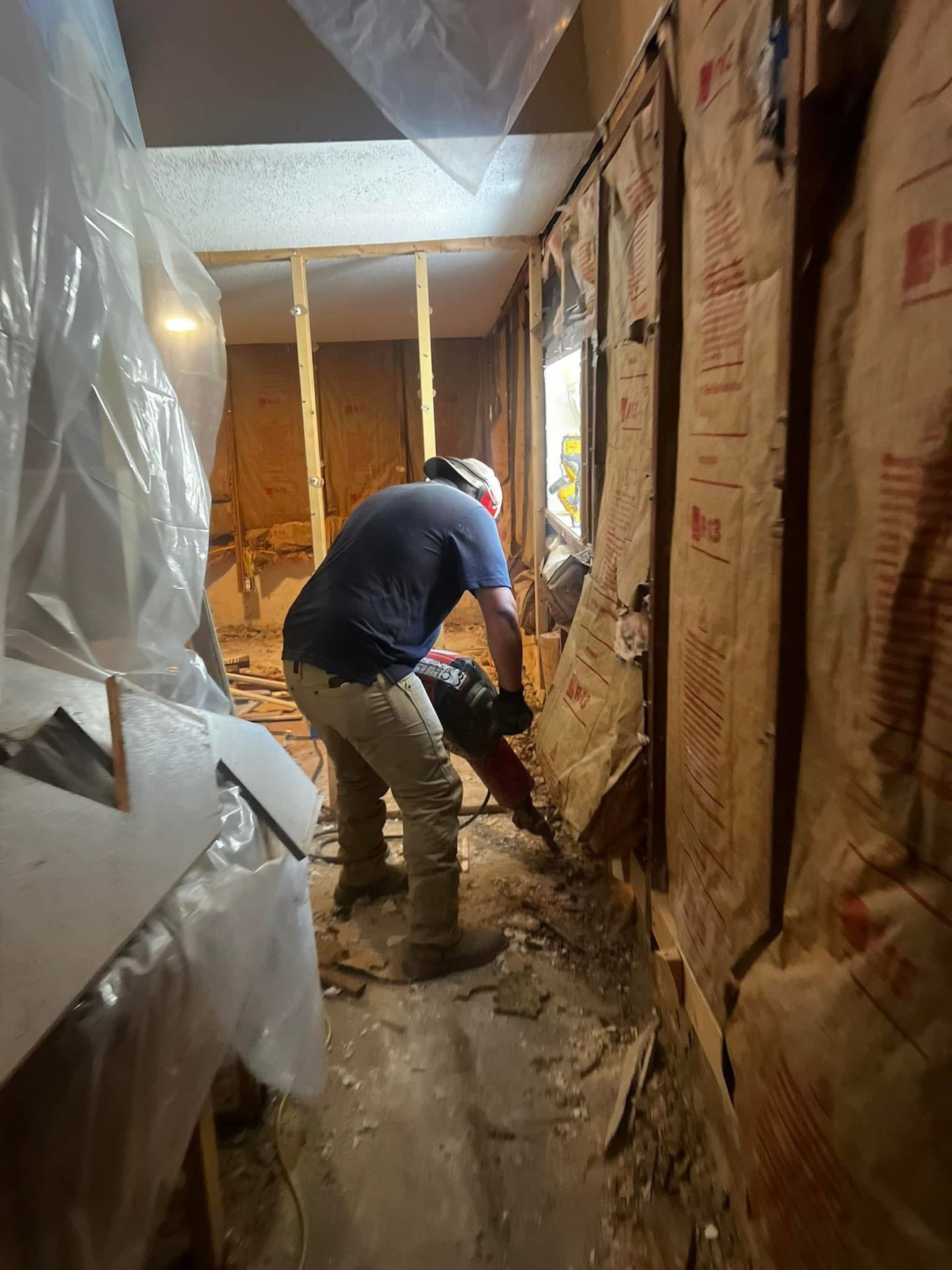 A worker in a blue shirt uses a power tool to break up a concrete floor in a room under renovation with exposed studs.