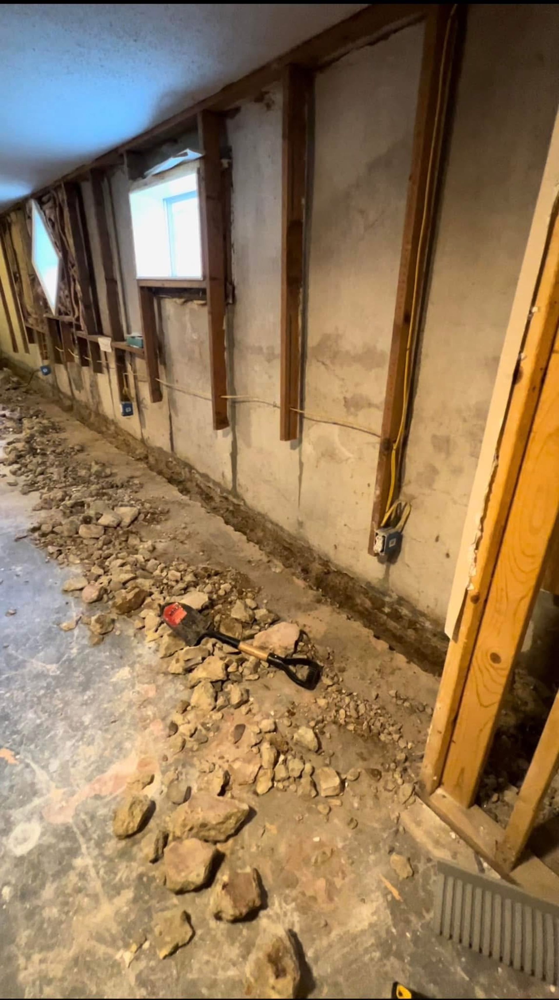 A basement wall with exposed wood studs, concrete foundation, and a trench of debris across the floor.