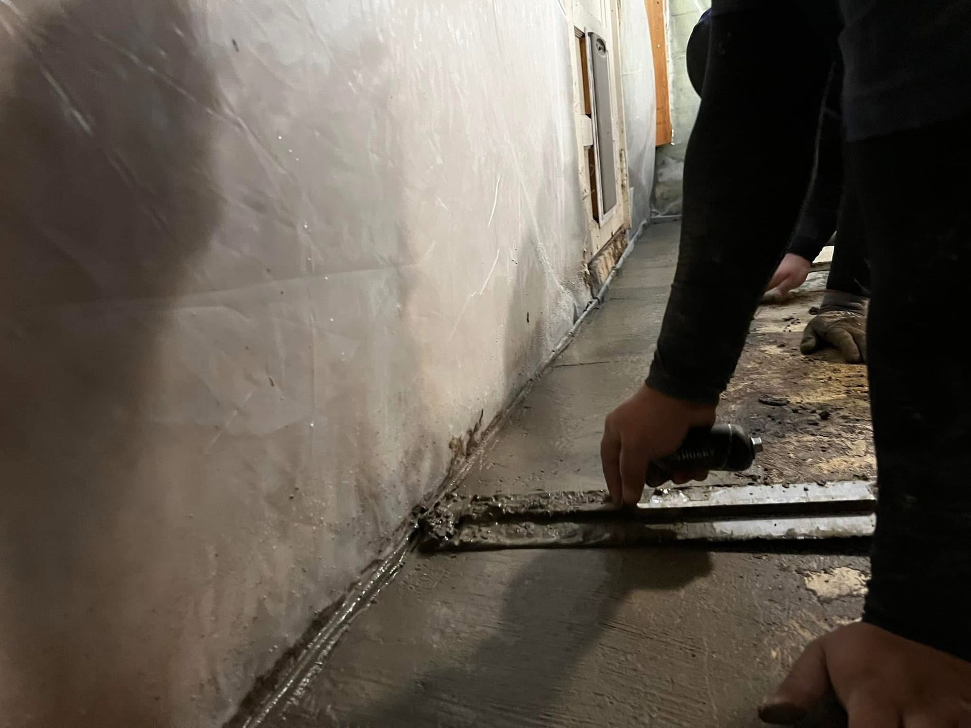 A close-up view of hands smoothing wet concrete along the base of a basement wall with a hand trowel.