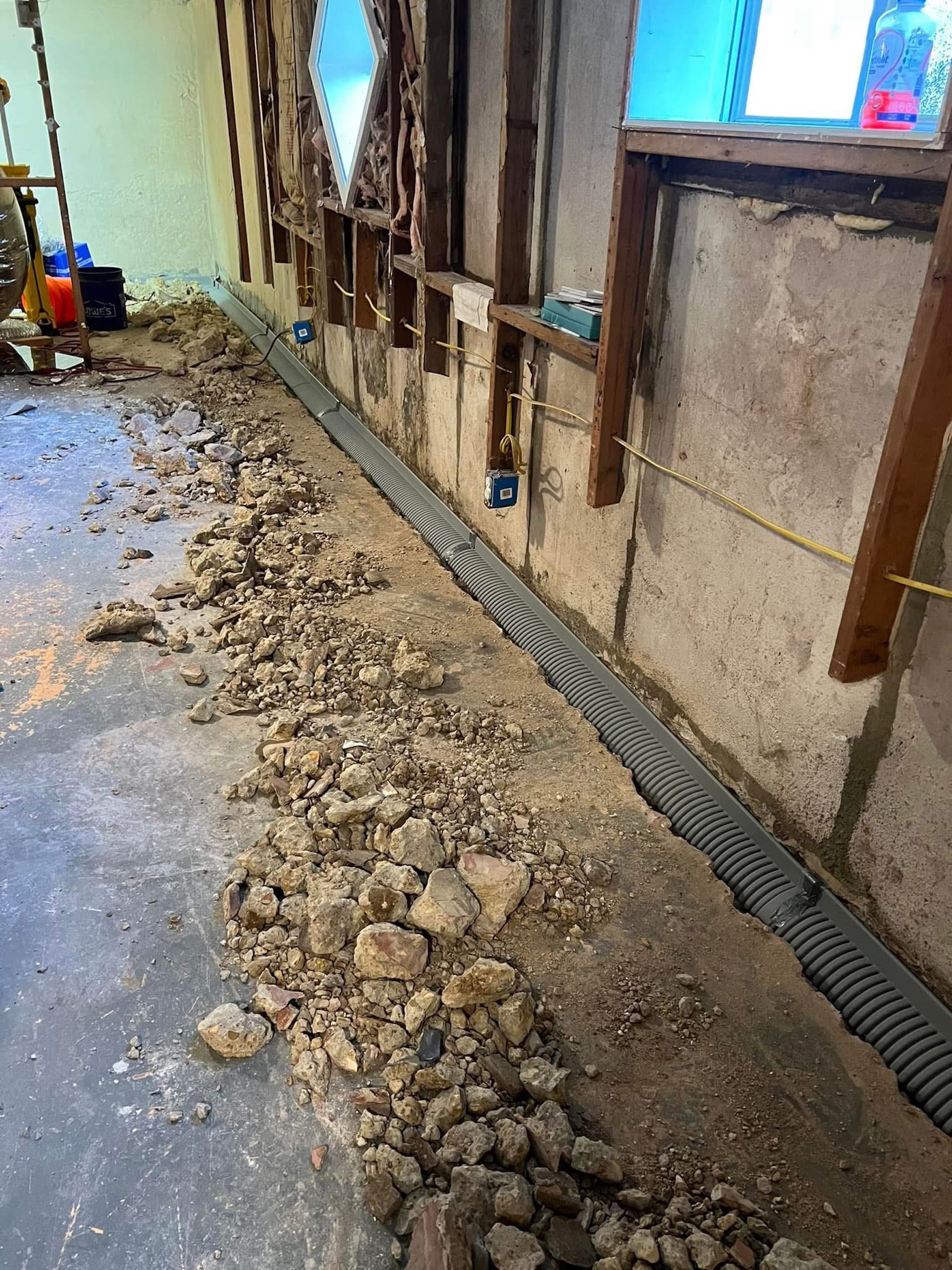 A basement interior under construction, showing a newly installed perimeter drainage pipe along an open wall foundation.