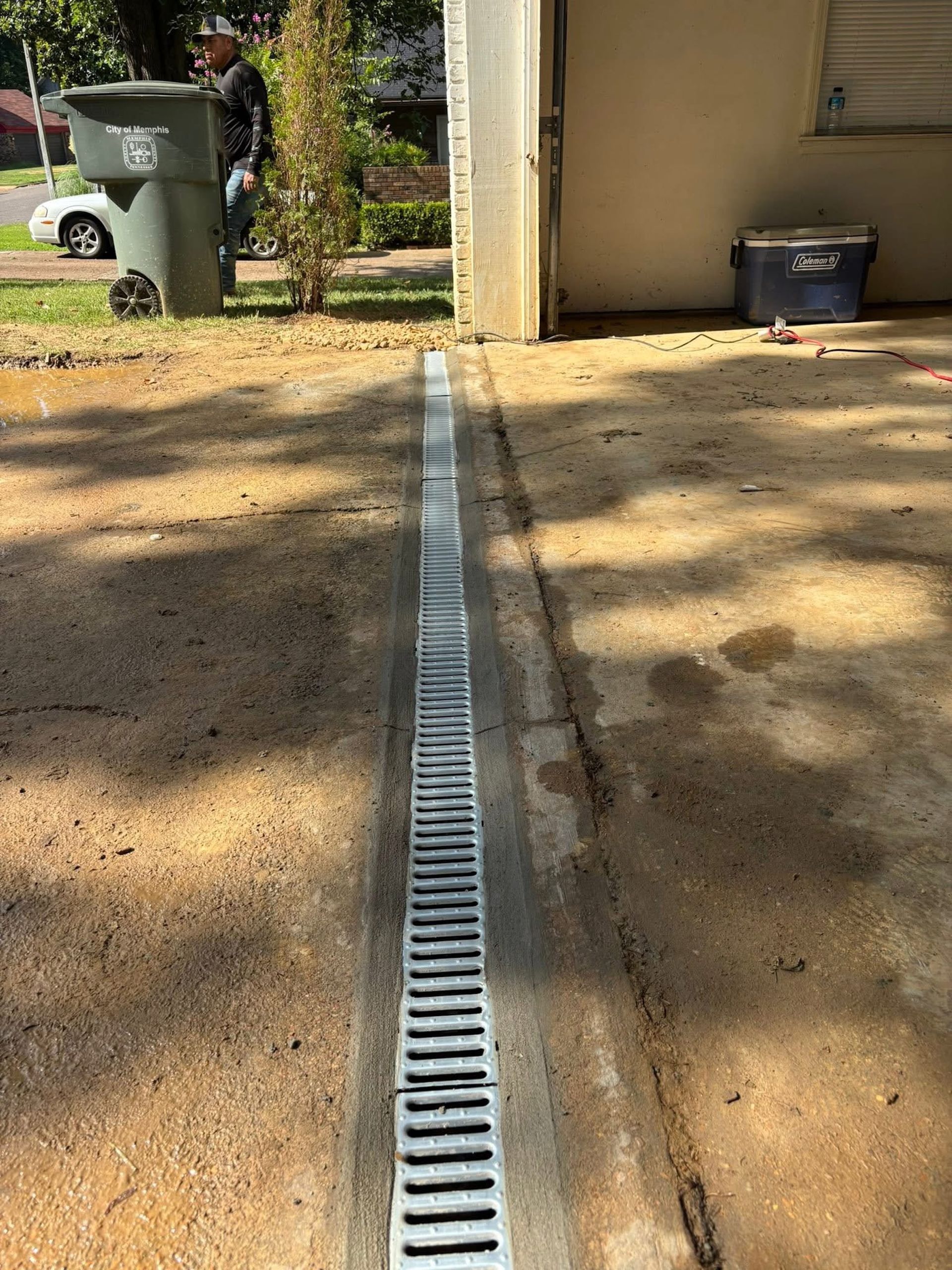 A metal trench drain grate embedded in a concrete driveway near a house exterior.
