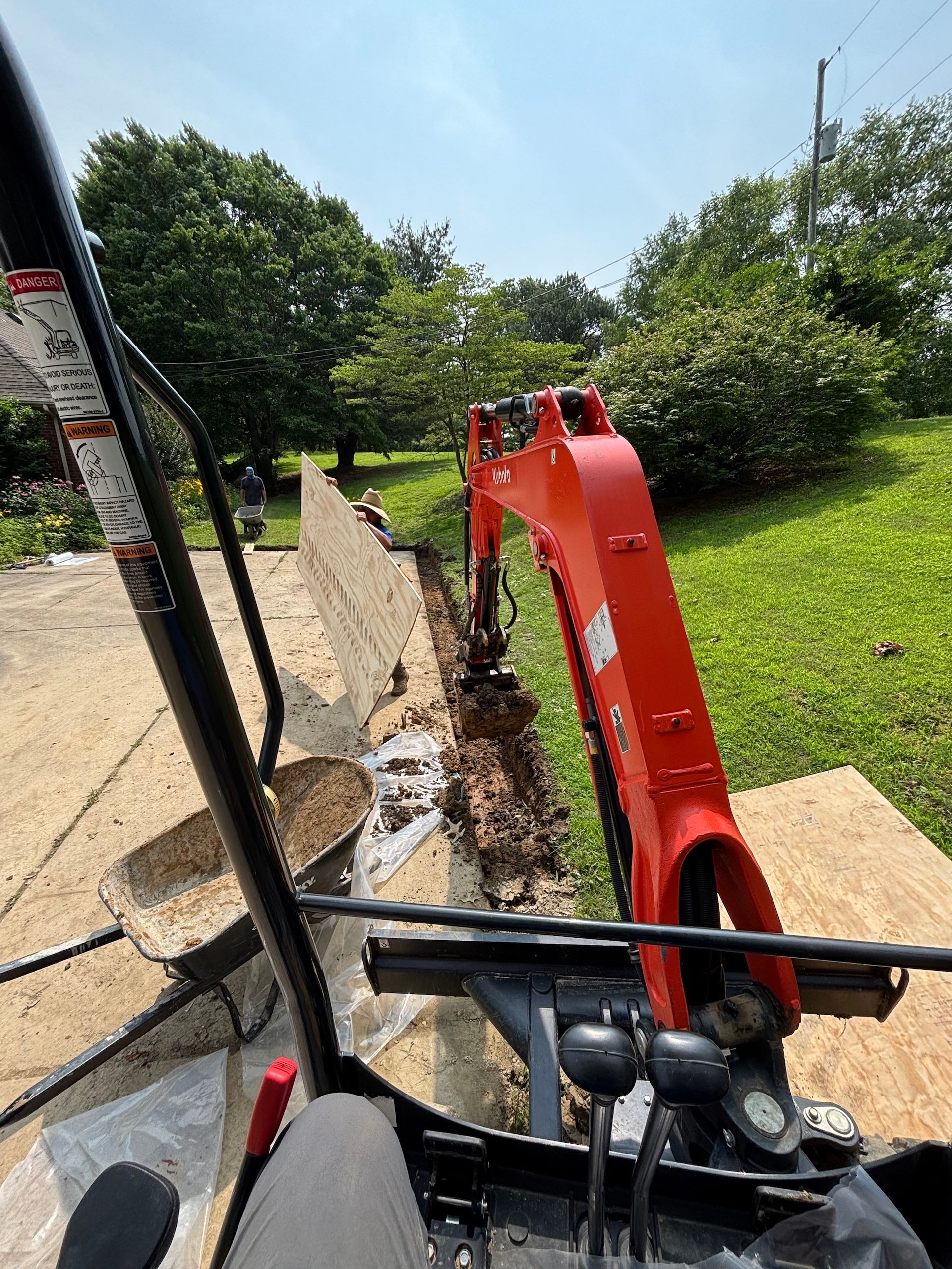 View from the cab of an orange excavator digging a trench along a low stone wall on a sunny, grassy lawn.