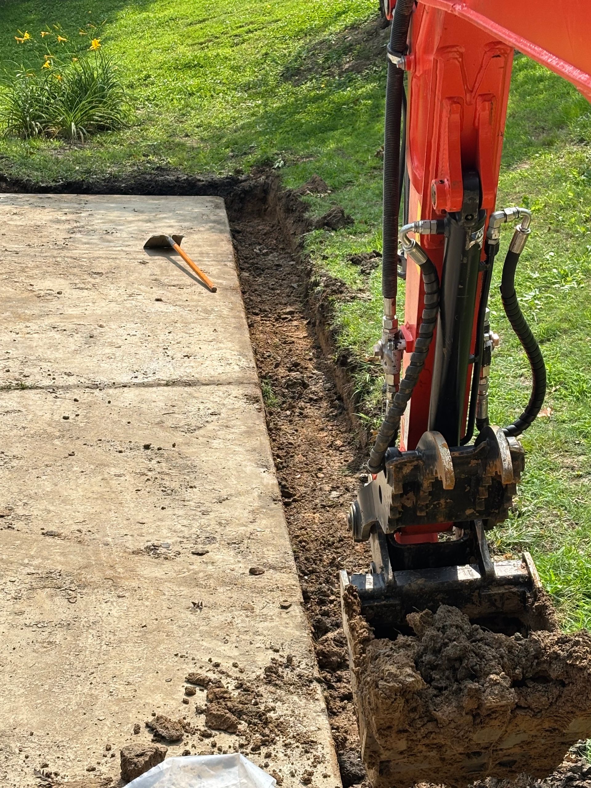 An orange mini excavator digging a trench along the edge of a concrete patio on a sunny, grassy day.