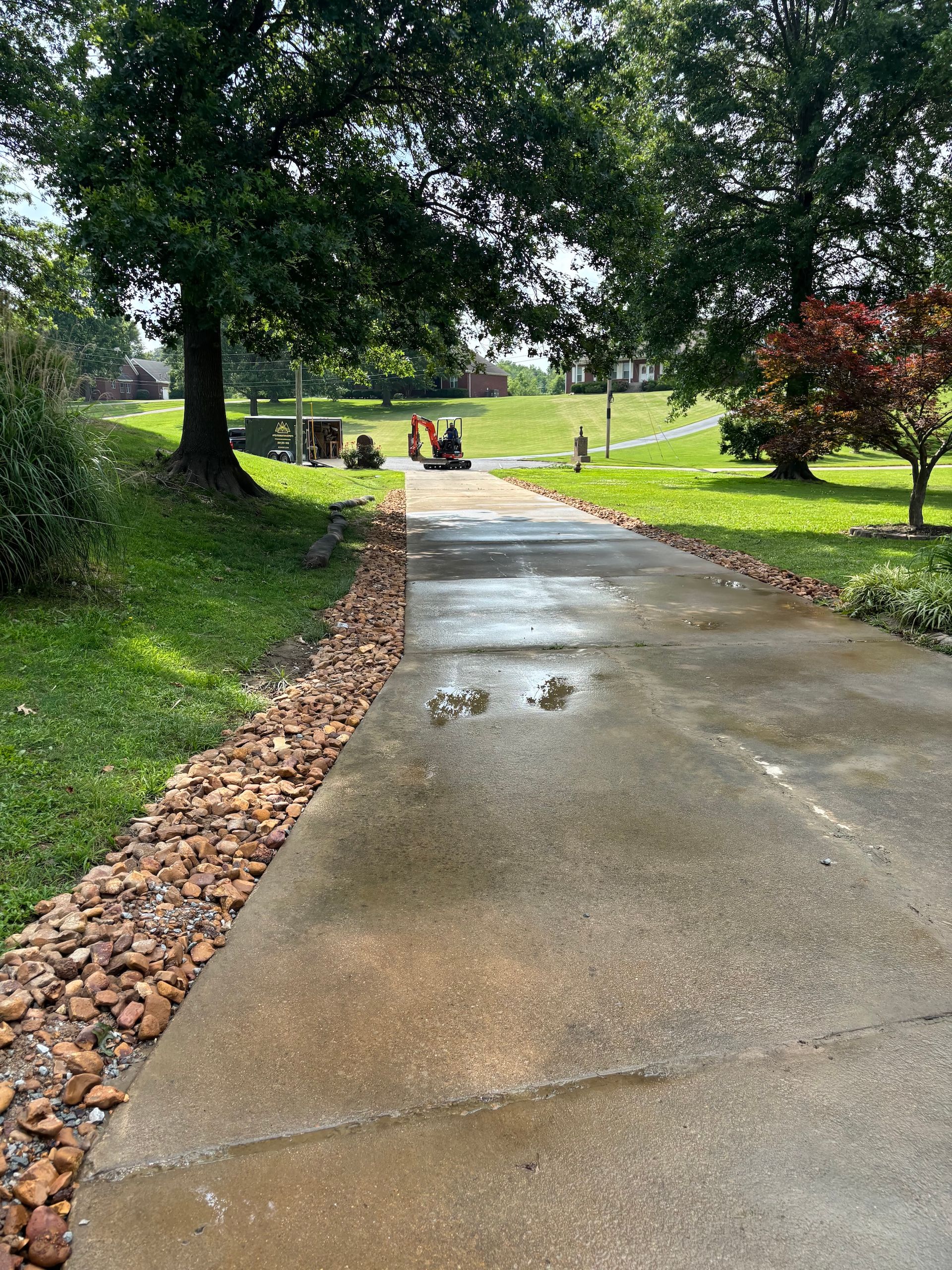 A wet, concrete driveway bordered by brown gravel, leading toward a grassy lawn under large shade trees.