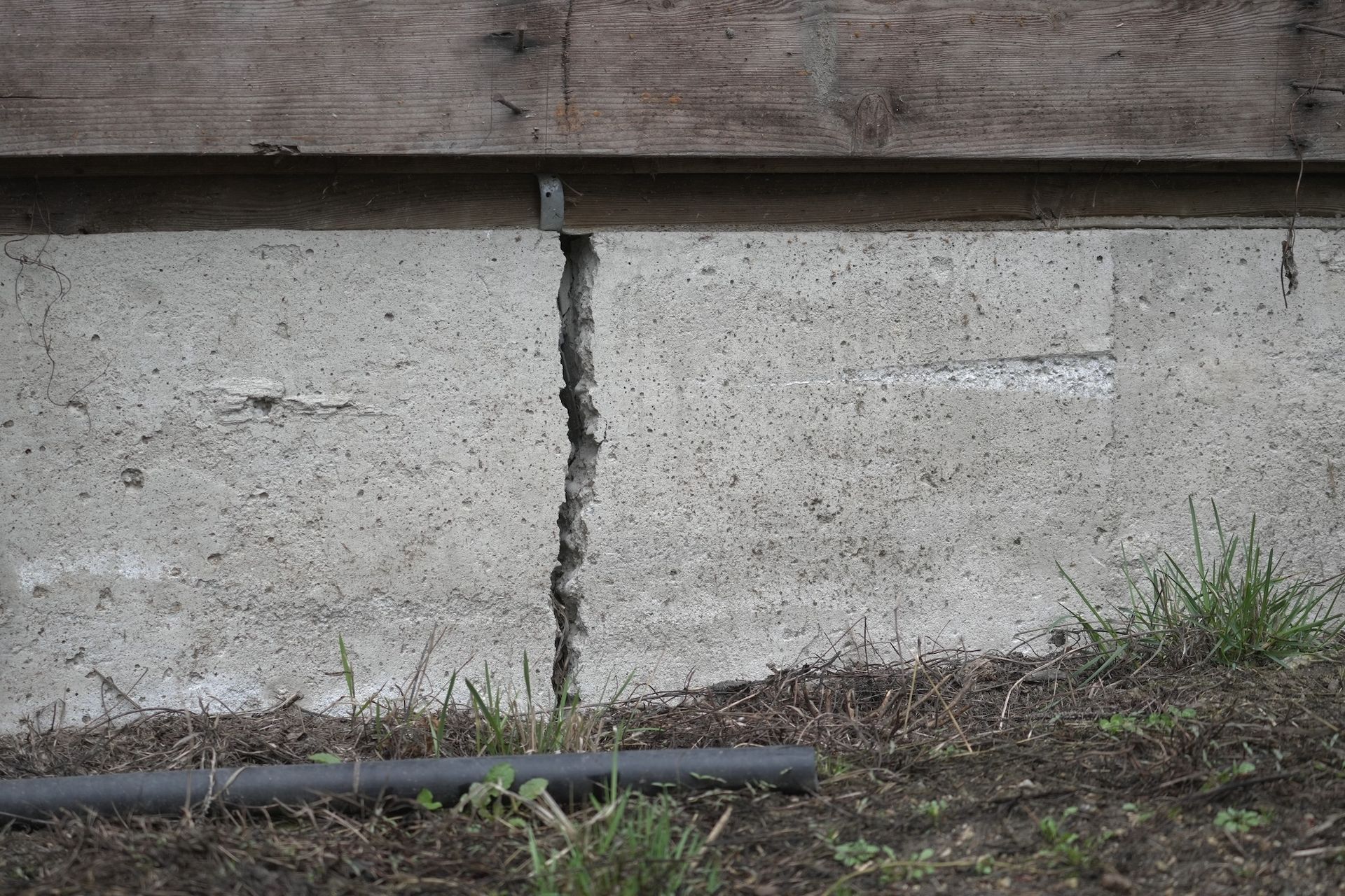 A vertical crack runs through a gray concrete foundation wall located beneath a wooden structure and soil.