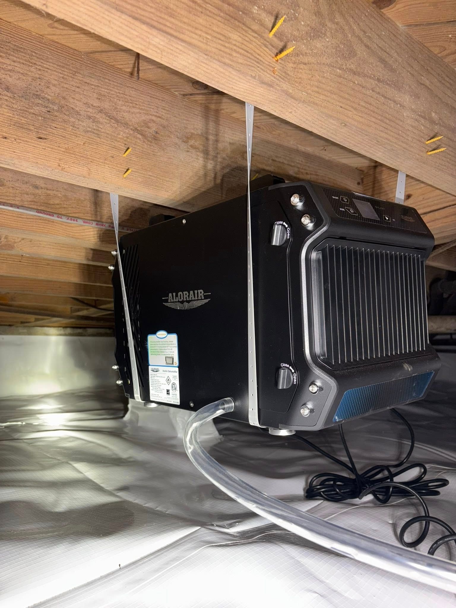 A black crawl space dehumidifier suspended from wooden floor joists above a white vapor barrier.
