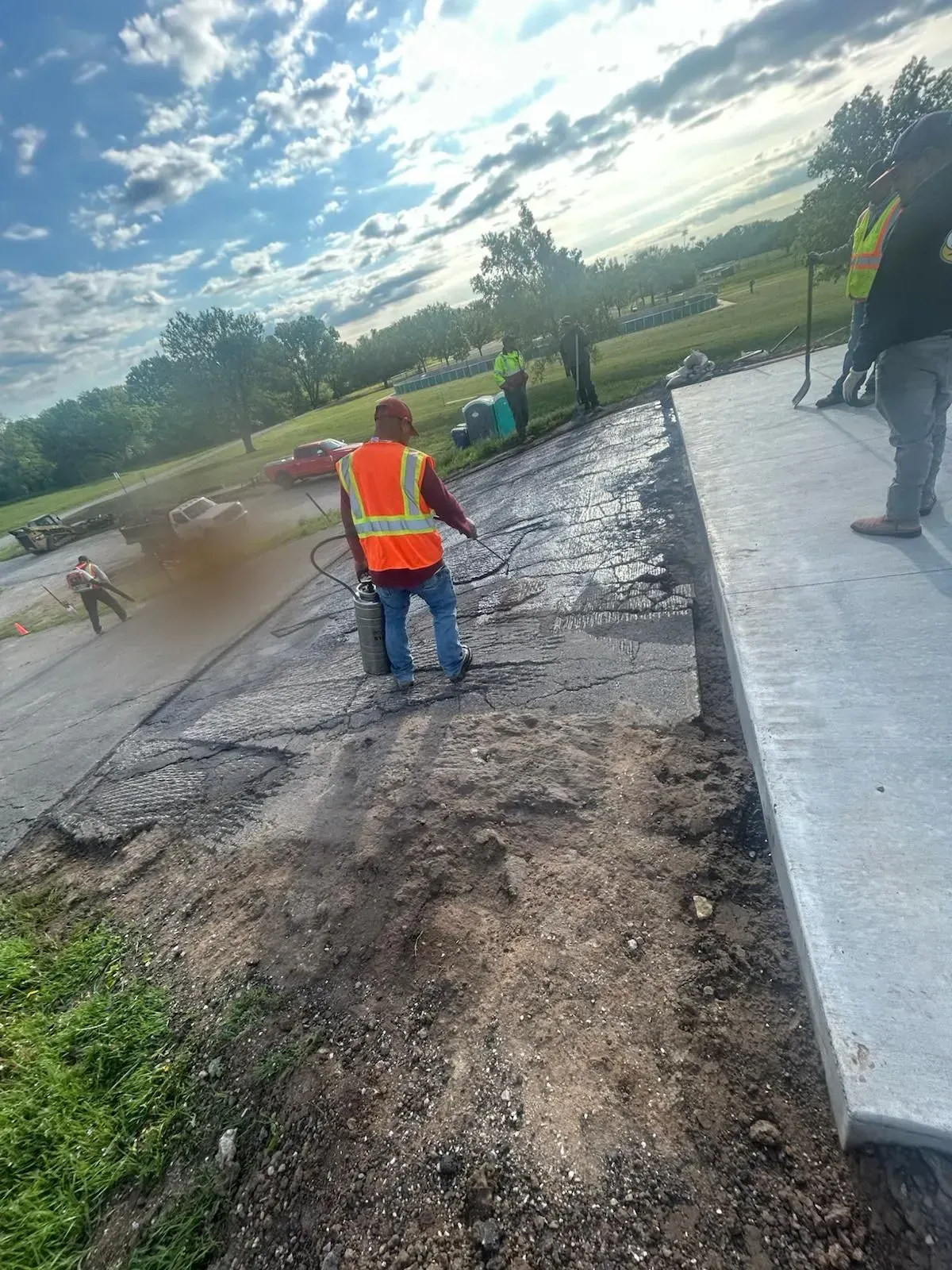Workers in orange vests paving a road beside a concrete curb at sunset