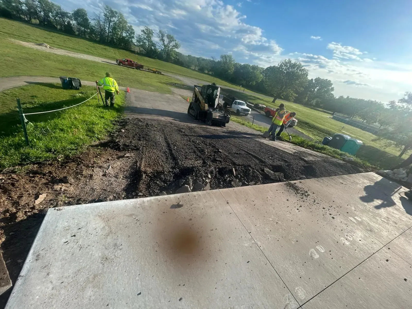 Workers and a small machine repair a damaged paved area beside a grassy roadside under a partly cloudy sky