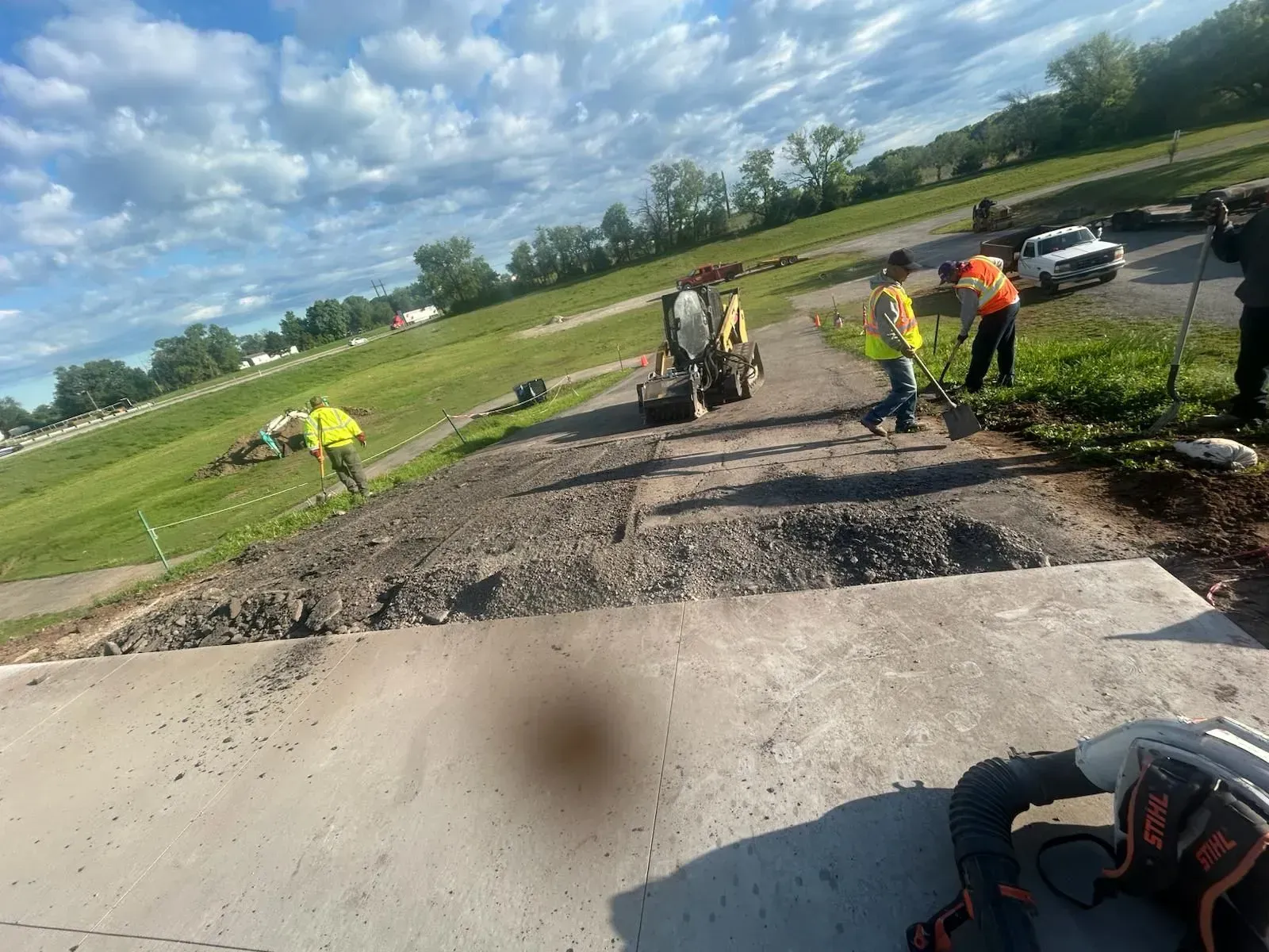 Roadwork crew repairing a damaged rural road with gravel and equipment under a cloudy sky