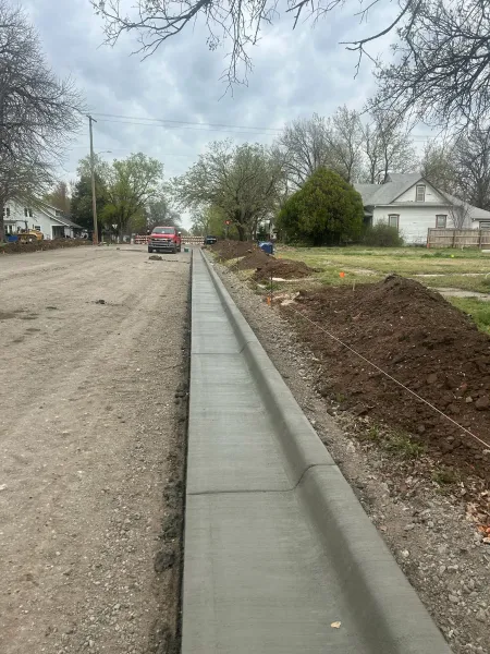 New concrete curb and gutter beside a gravel street under construction, with trees and houses nearby