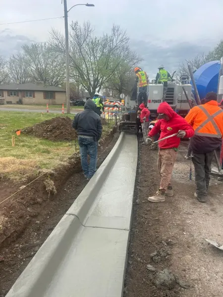 Workers installing concrete curb along a trench on a residential street, with one person in a red jacket supervising.