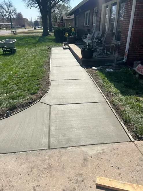 Concrete walkway beside a brick house, leading through a grassy yard on a sunny day.