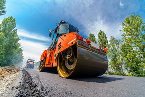Orange road roller paving asphalt on a road under blue sky, low angle view with trees nearby