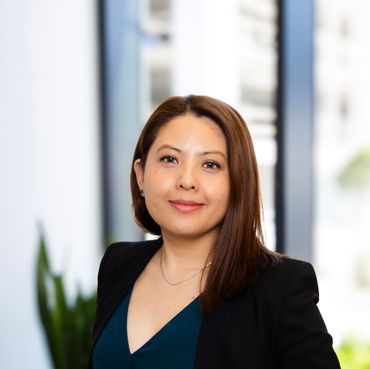 Portrait of a woman in a black blazer and teal top, standing indoors with a blurred office background