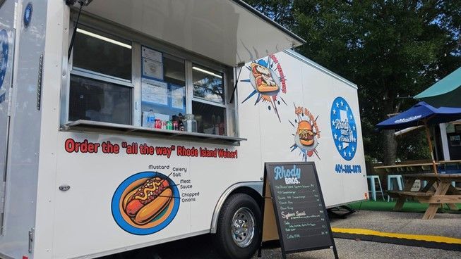 A white food truck with a wiener on the side is parked in a parking lot.