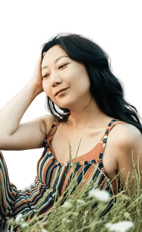 Woman in striped jumpsuit, hand in hair, sitting in tall grass, looking relaxed.
