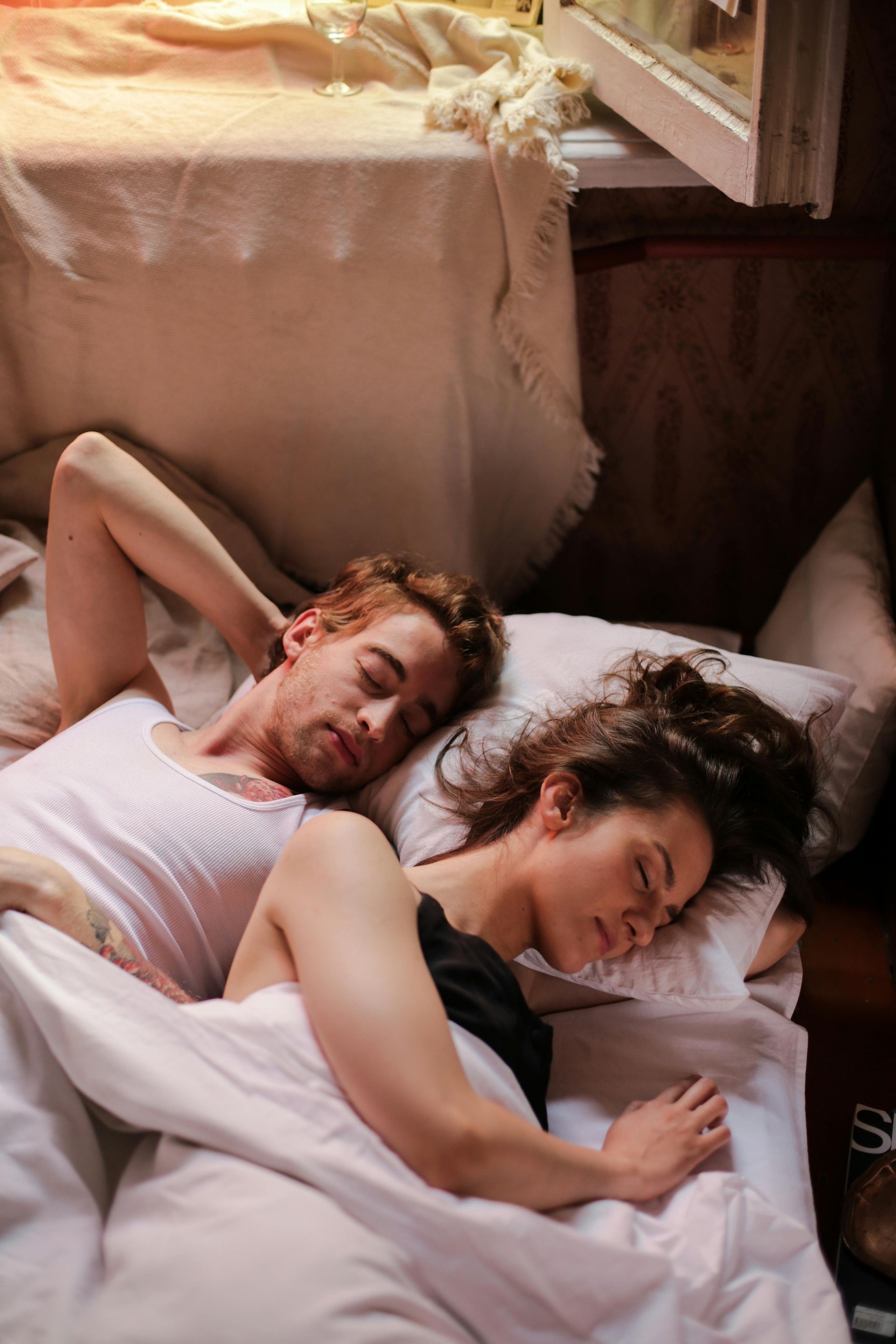 Couple asleep in bed, covered with a white blanket. Warm light and cozy setting.