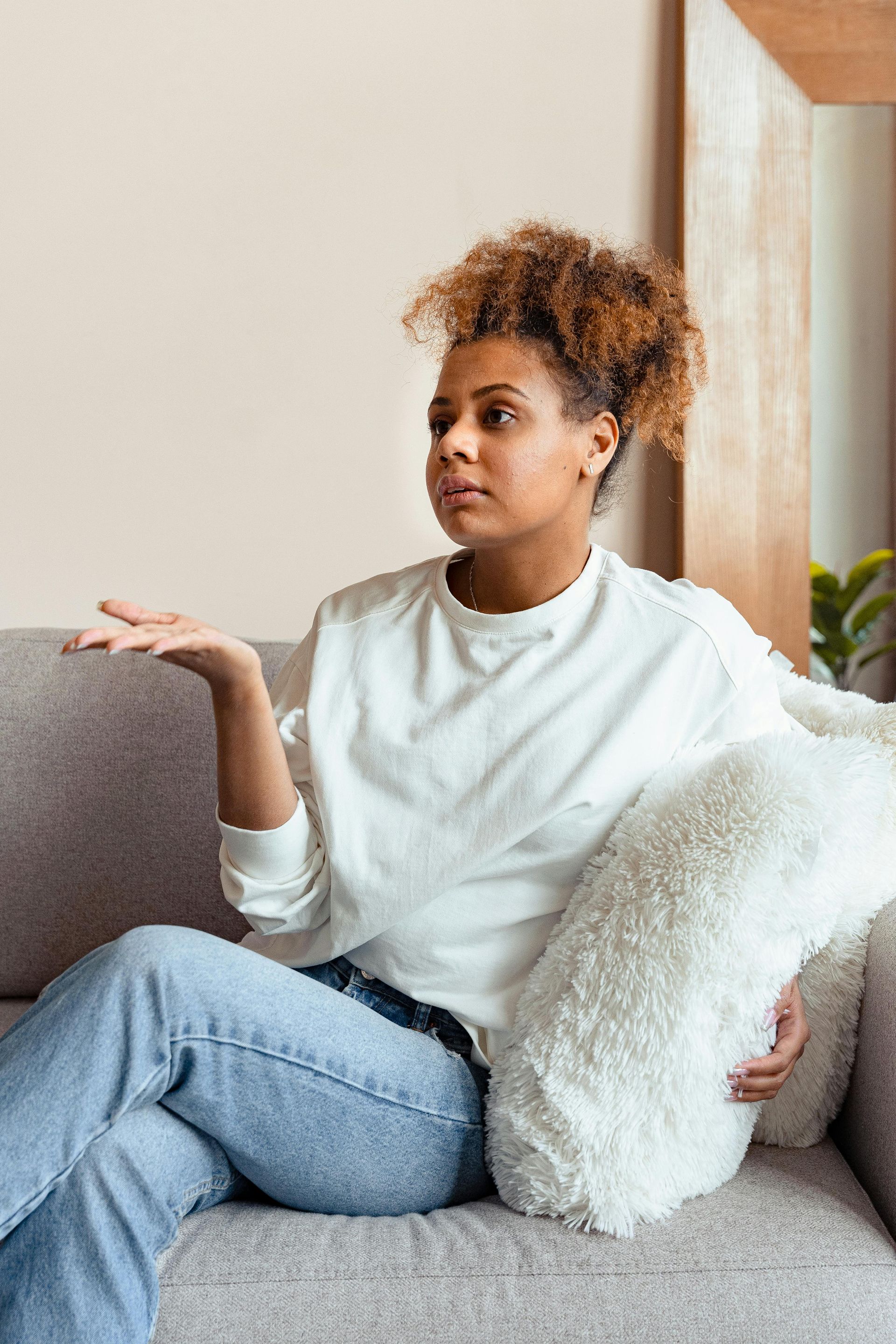 A person with curly hair sits on a gray couch, wearing a white sweater and jeans, holding a fluffy pillow while gesturing.