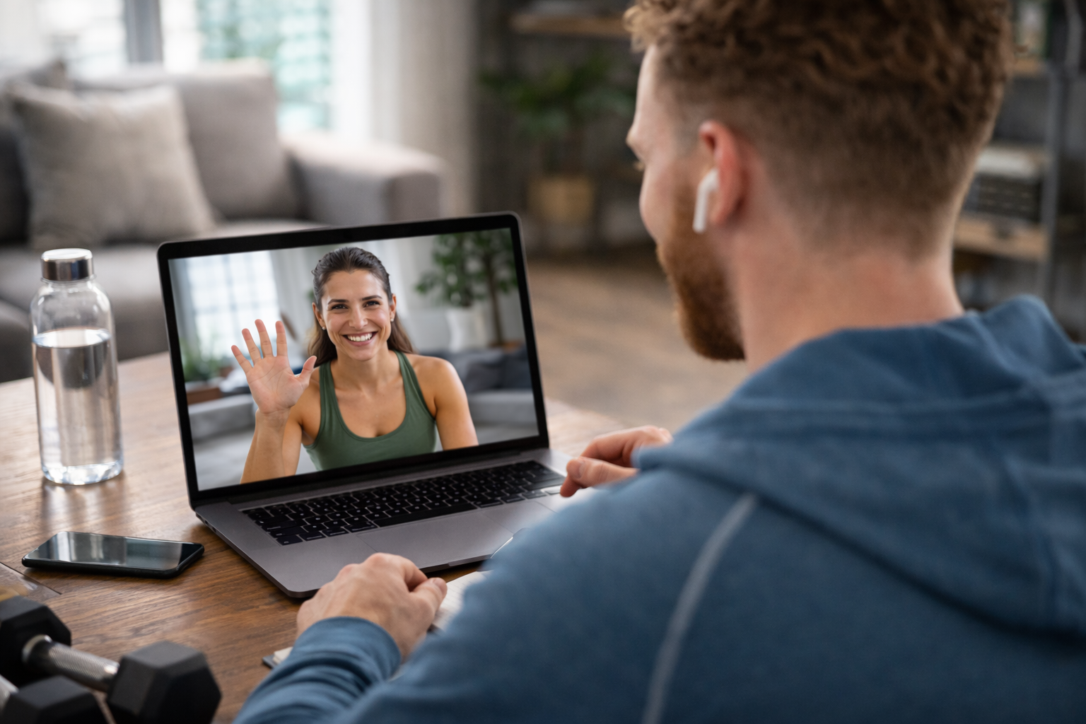 Iemand die via een laptop videobelt, zwaait vanaf de bank met een fles water naast zich.