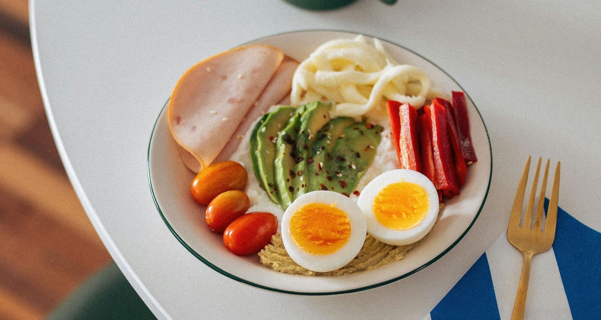 Een bord met eten met eieren, ham, avocado, tomaten en paprika's op een tafel.