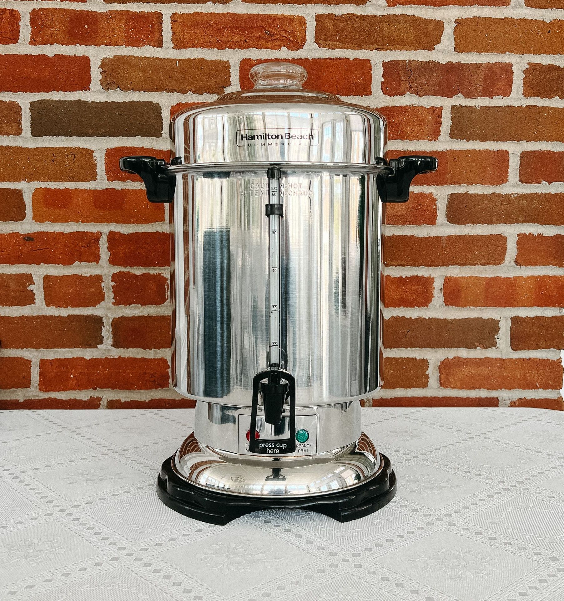 A stainless steel coffee percolator is sitting in front of a brick wall