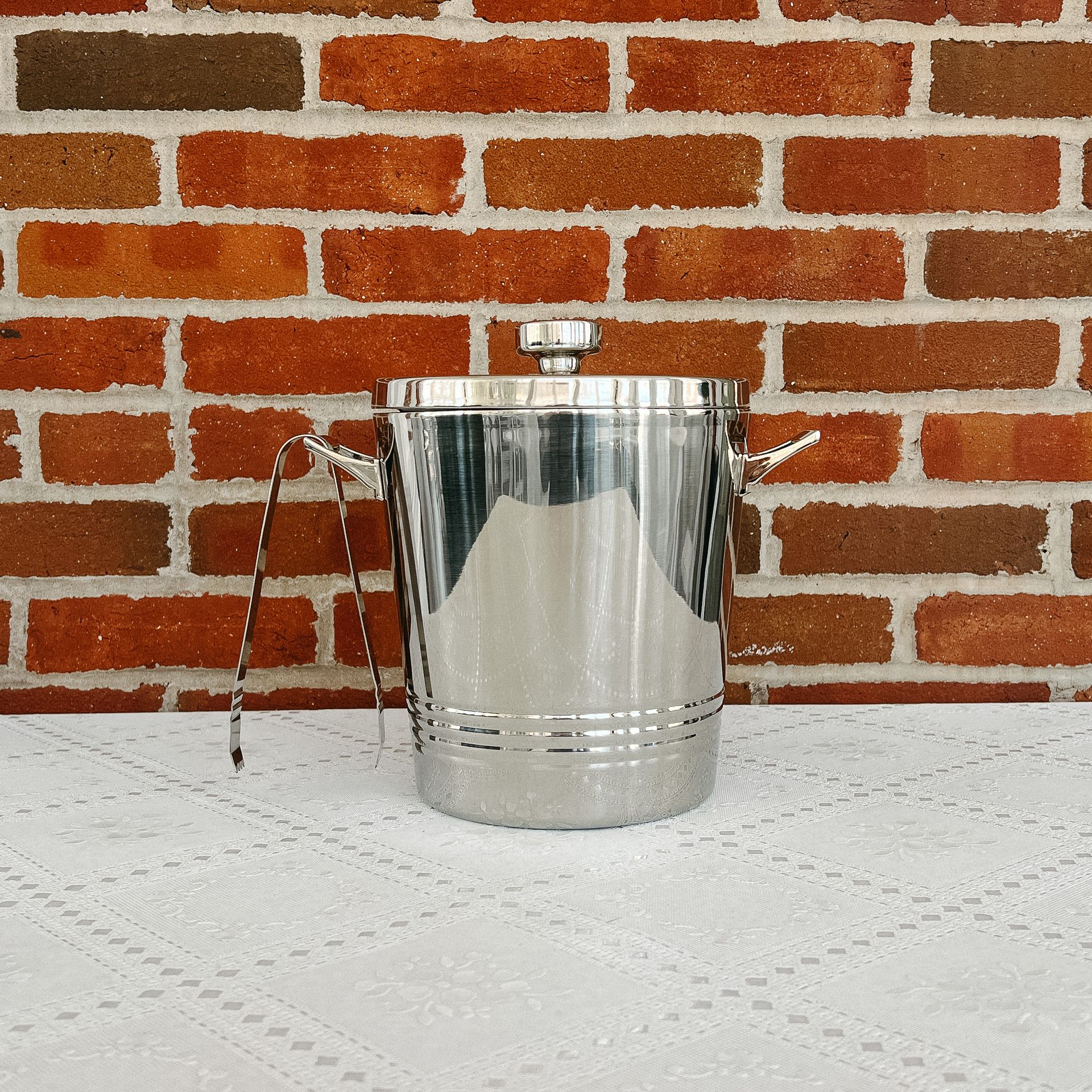A stainless steel ice bucket sits on a table in front of a brick wall