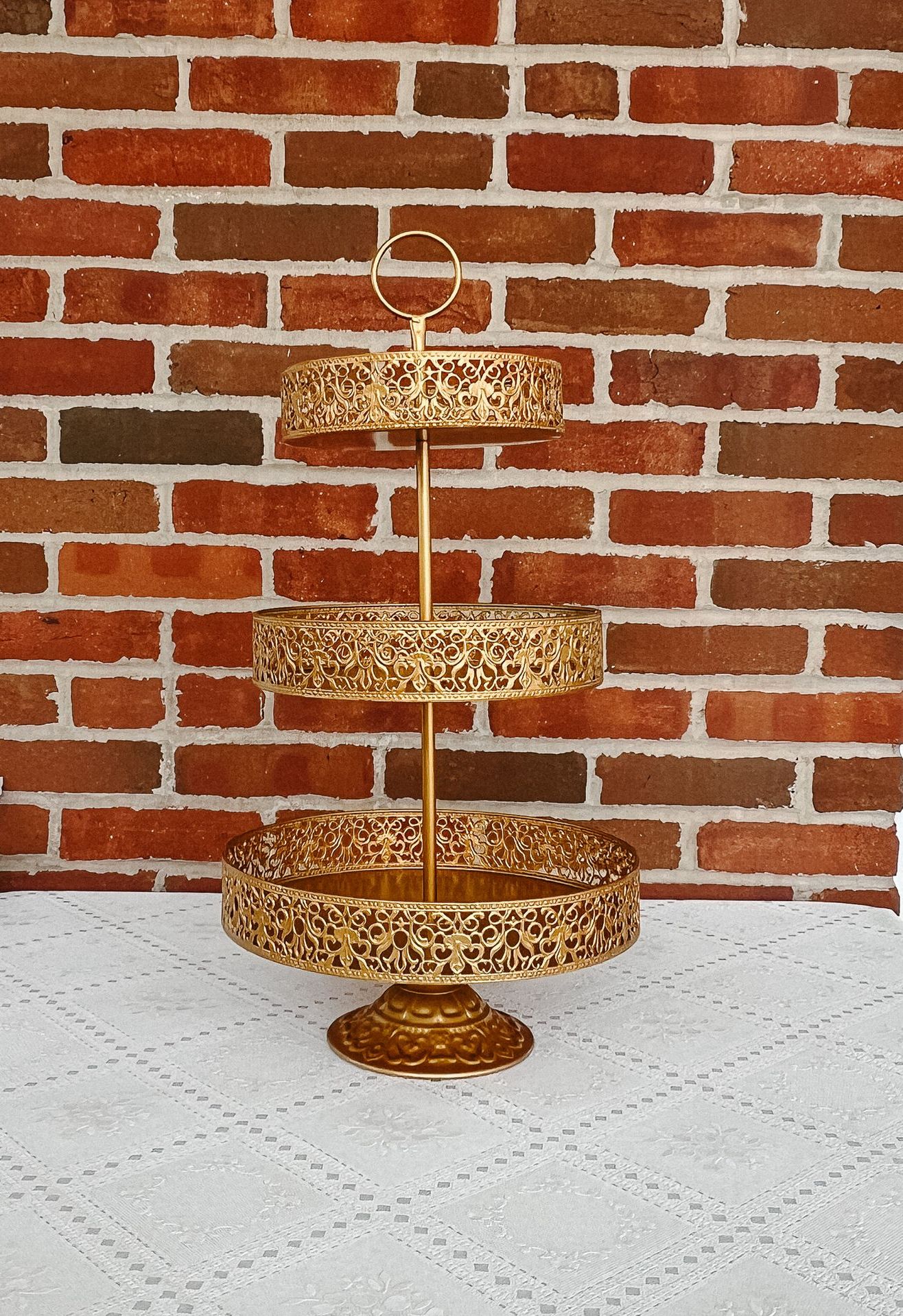 A gold cake stand is sitting on a table in front of a brick wall.