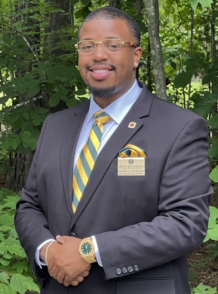 Professional portrait of a person in a dark suit, green and gold tie, and name tag, standing outdoors with trees.