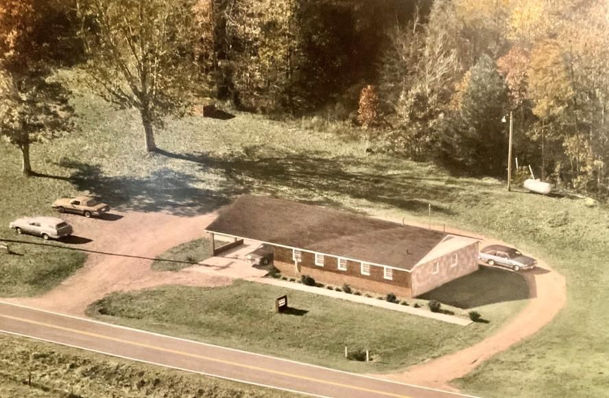 An aerial view of a rectangular, single-story building with a brown roof, situated next to a road with parked cars nearby.