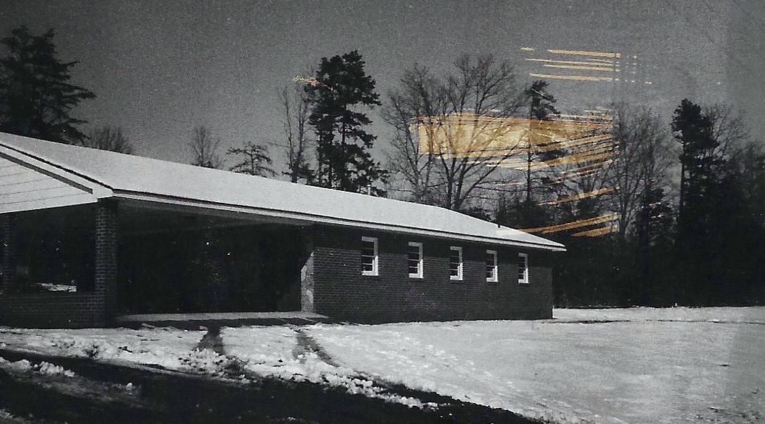 A single-story building with a flat roof and side windows sits in a snow-covered field with trees in the background.