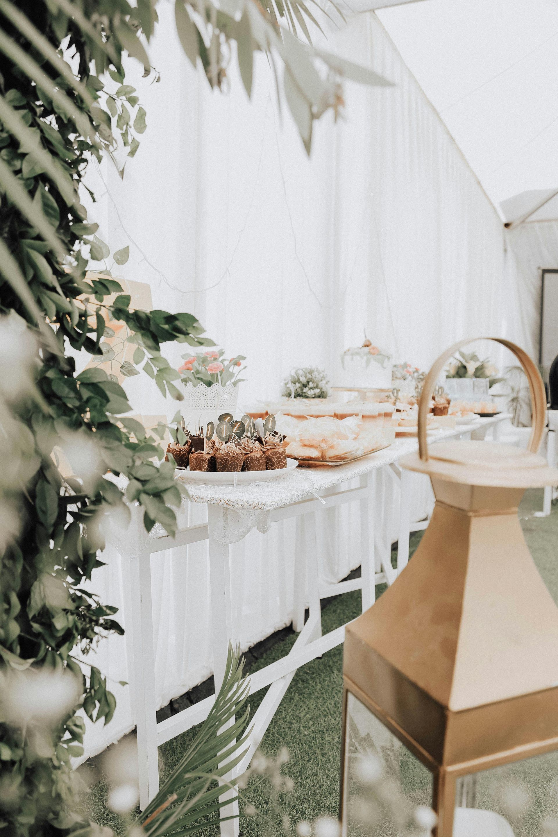 Buffet table with desserts, greenery, and gold lantern in a white tent setting.