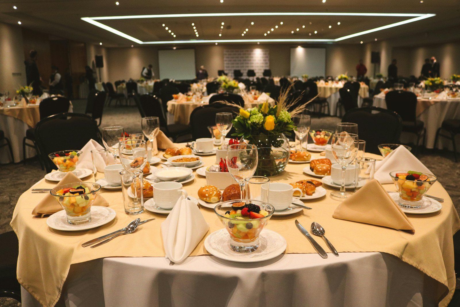 Round tables set for a formal event with fruit salads, drinks, and flower centerpieces in a ballroom.