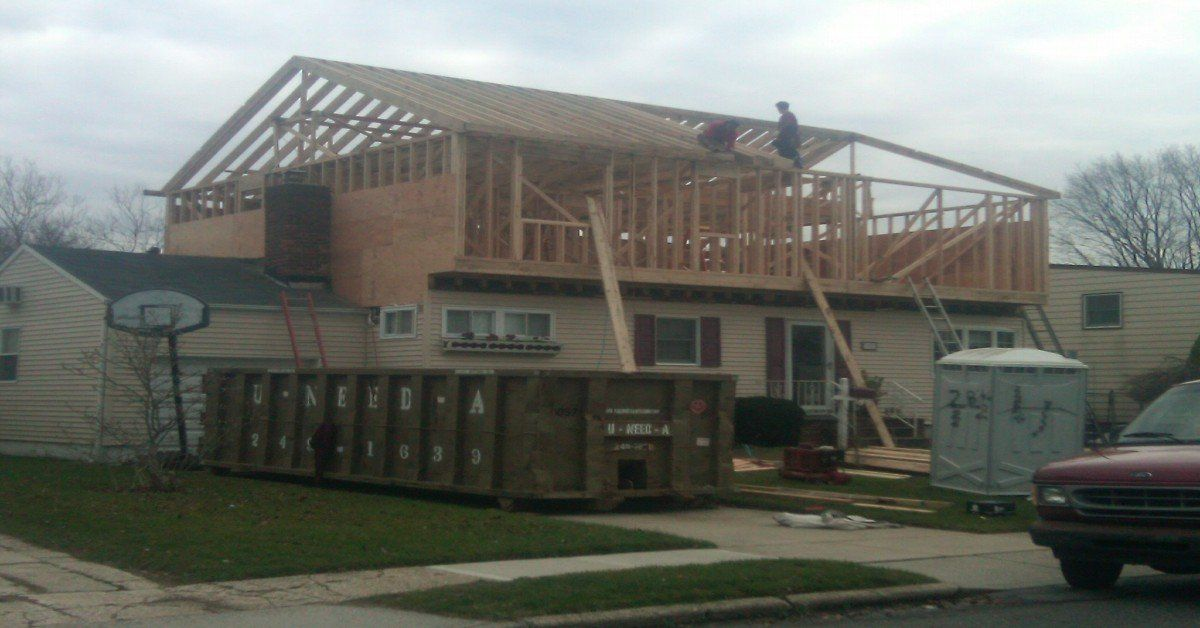 House under construction with workers on the upper frame, a dumpster, and a portable toilet in front.