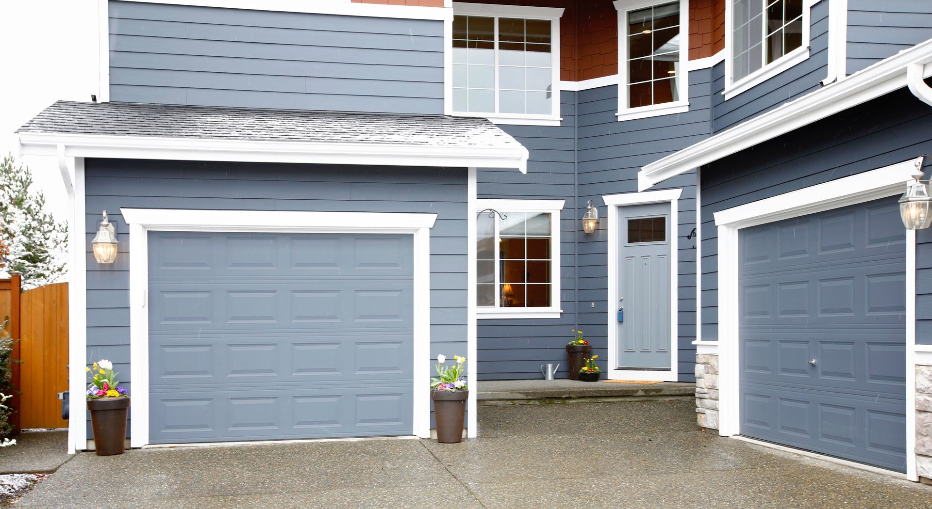 Blue-sided house with two garages, a front door, and white trim. Gray roof and concrete driveway.
