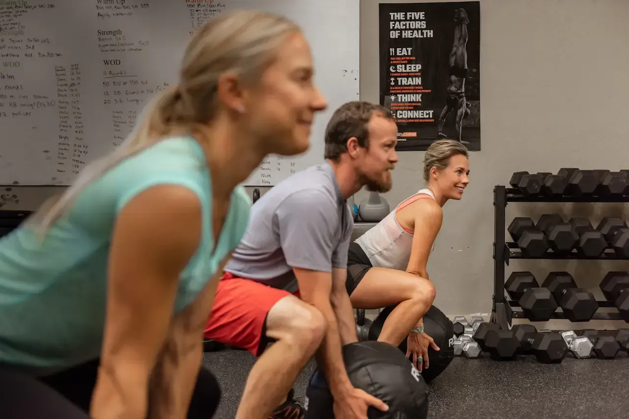 Three people in a gym doing squats with medicine balls. Gray wall, dark dumbbells.