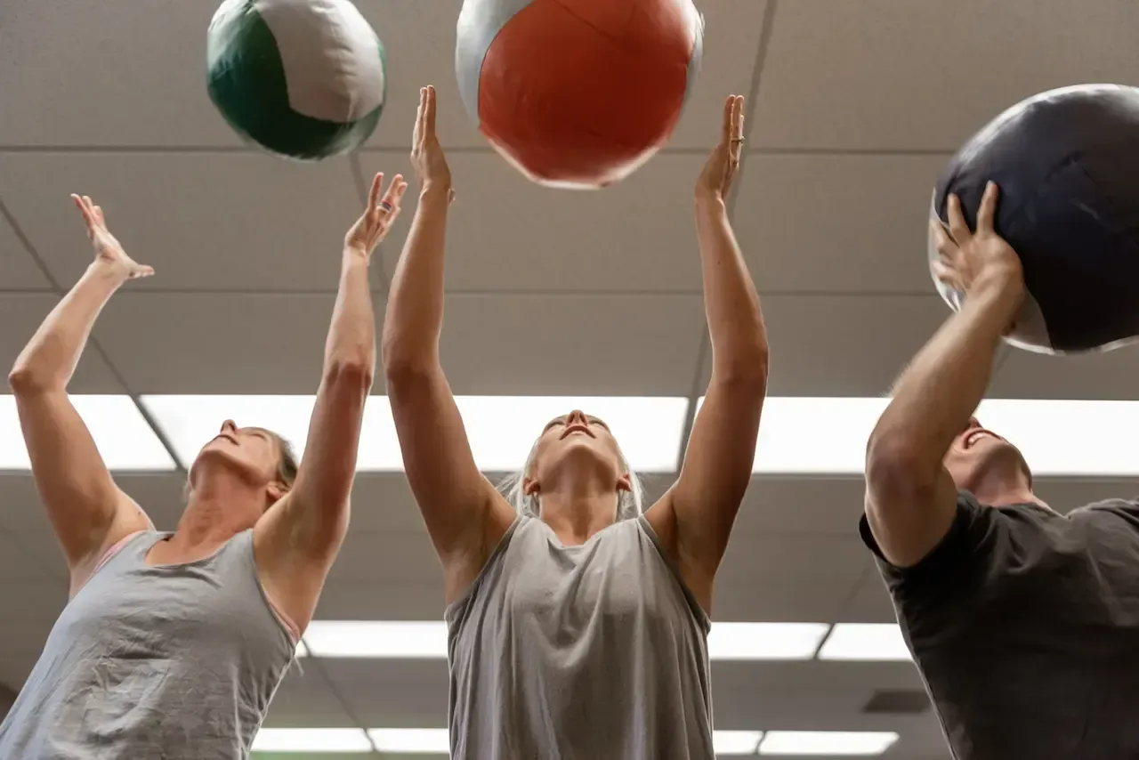 Three people reaching upwards to catch exercise balls in a gym.