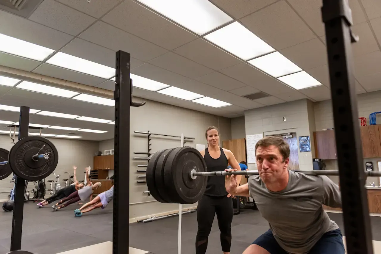 Man squats with barbell, assisted by woman, in gym. Others exercise in background.