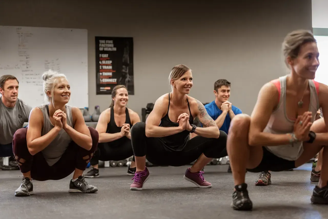 People in a gym, squatting low, hands together, smiling. Gray walls, weights, a white board.