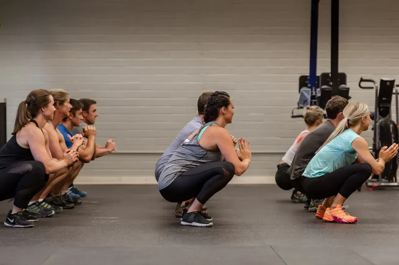 People squatting in a gym, arms folded, feet flat on the floor, in a group exercise setting.