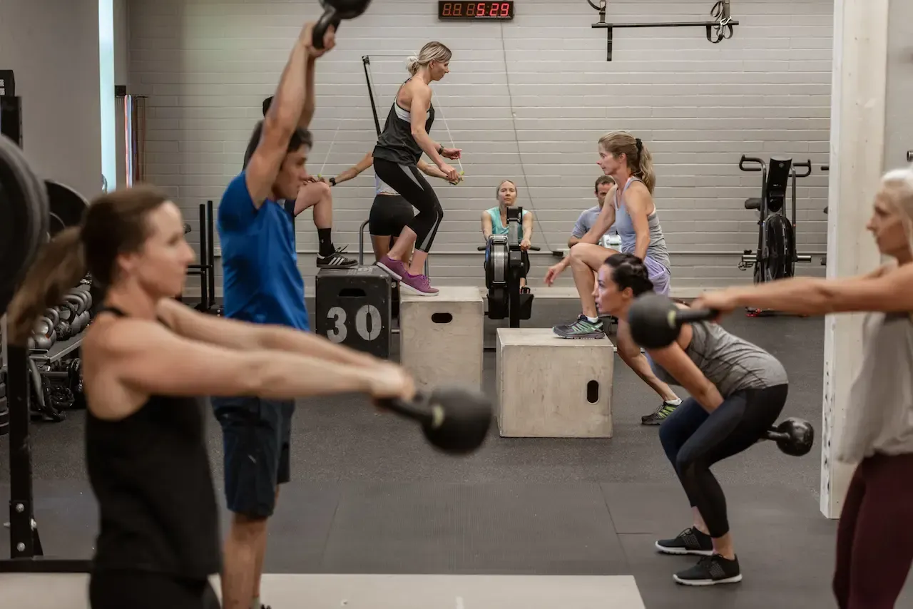People exercising at a gym; kettlebell swings, box jumps, rowing machine, neutral setting.