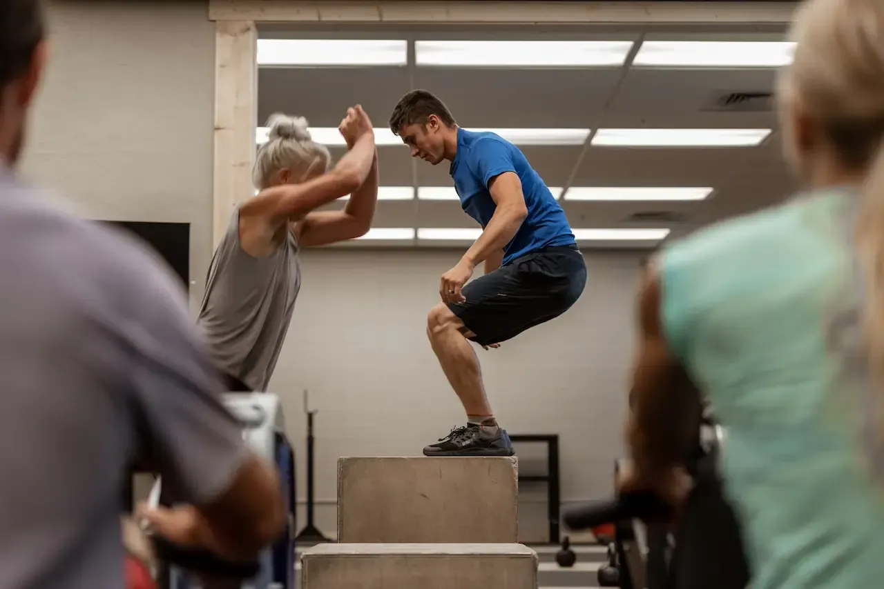 Man jumping onto a box, with a woman assisting him in a gym.