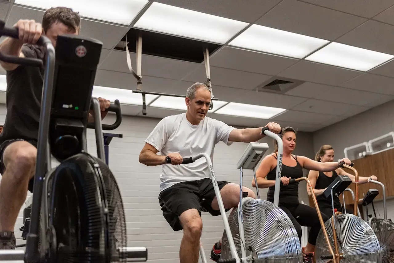 People exercising on air bikes in a gym. Man in white shirt smiles, others focused. Bright overhead lighting.