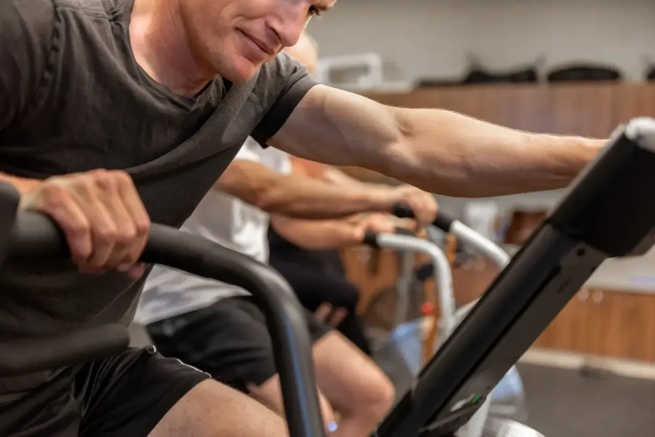 Man exercising on a stationary bike in a gym, arm extended to hold the handlebars. Another person is visible in the background.